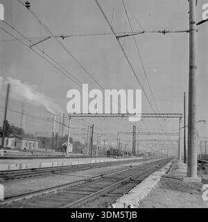 Slawjansk, Region Donezk, ukrainische SSR, UdSSR. 1970er Jahre Eine atmosphärische Archivansicht der Bahnhofsgleise, die sich bis in den industriellen Horizont erstrecken. Das komplexe Netz von Freileitungen hängt über den Schienen. In der Ferne dominieren hohe Rauchschornsteine eines Wärmekraftwerks oder einer Fabrik die Skyline und betonen die Schwerindustrie der Region. Rechts steht ein Betonpfosten im Vordergrund. Das Bild erfasst die Größe der Verkehrsinfrastruktur, die Industrielandschaft und die trübe Atmosphäre des friedlichen Donbass. Stockfoto