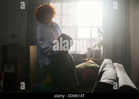 Kaukasische Frau mittleren Alters stehend und sanft mit dem Gesicht eines jungen Erwachsenen kaukasischen Mannes, der auf der Couch sitzt. Beide teilen sich liebevolle Momente im sanft beleuchteten Wohnzimmer Stockfoto