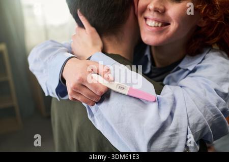 Junge kaukasische Frau lächelt, während sie den jungen Mann umarmt, einen positiven Schwangerschaftstest in der Hand hält, beide stehen drinnen und drücken Glück und Aufregung über die Schwangerschaftsnachrichten aus Stockfoto