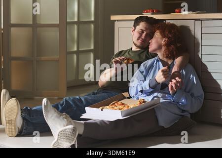 Junge Liebhaber sitzen auf dem Küchenboden und essen gemeinsam Pizza, lächeln und lehnen sich nahe, teilen intime Momente mit offener Pizzabox Stockfoto