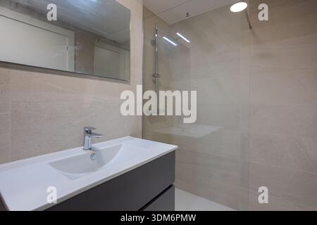 Modernes Bad mit beigefarbenen Wänden, begehbarer Glasdusche, Spiegel und elegantem Kosmetikbereich Stockfoto
