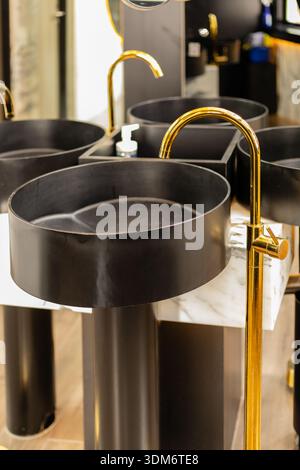 In einem modernen Badezimmer stehen mehrere schwarze runde Waschbecken. Die Waschbecken haben goldene Armaturen und einen Marmorsockel. Das Gesamtbild ist sauber und elegant Stockfoto