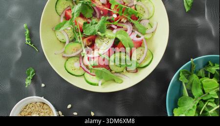 Blassgelbe Salatschüssel auf dunkler Matte mit Gurke, Rucola, Samen; weiß, blaue Schüsseln Stockfoto