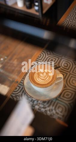 Flacher weißer Kaffee mit Latte Art auf einem gefliesten Holztisch in einem unabhängigen Café in Großbritannien. Natürliches Licht, handwerkliches Café-Ambiente Stockfoto