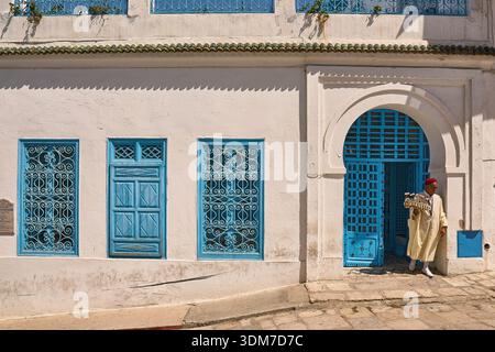 Mann im traditionellen tunesischen Kaftan in der Nähe der hellen blauen Tür und der verzierten Fenster in der Sunny Street in Sidi Bou Said Stockfoto