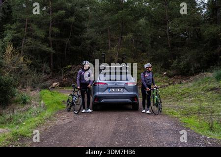 Porträt von zwei Radfahrern mit passenden Trikots und Helmen, die auf beiden Seiten eines grauen Toyota Corolla Cross Hybrid-SUV stehen. Stockfoto