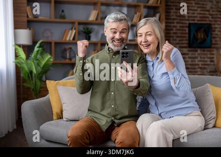 Ein aufgeregtes Seniorenpaar sitzt auf einem Sofa, schaut auf einen Smartphone-Bildschirm, jubelt und drückt Freude aus, feiert einen Sieg oder erhält dank moderner digitaler Technologie gute Nachrichten Stockfoto