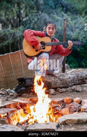 Frau spielt Akustikgitarre am Lagerfeuer, während sie draußen campt Stockfoto