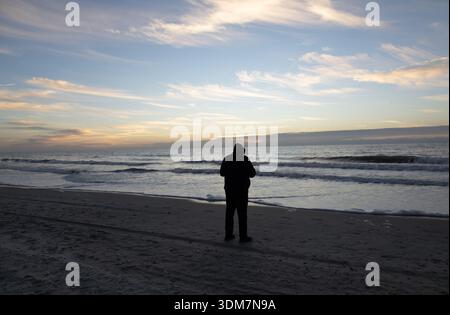 Silhouette einer Person, die bei Sonnenaufgang an einem Meeresstrand steht und in Richtung Horizont blickt, mit ruhigen Wellen und sanftem Morgenlicht. Stockfoto