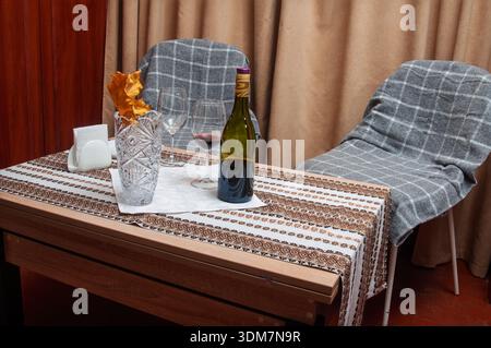 Eine Flasche Rotwein und zwei Gläser stehen auf einem Tisch mit einem dekorativen Läufer neben einer Kristallvase, die getrocknete Blätter enthält, und sorgen für eine einladende Atmosphäre Stockfoto