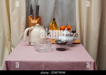 Auf einer rosafarbenen Tischdecke befinden sich eine Sammlung von Küchenartikeln, darunter eine Teekanne, ein Wasserkocher, eine Obstschale und ein Messerblock Stockfoto
