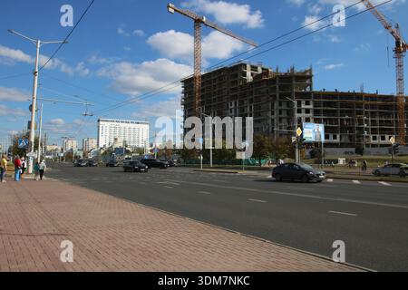 Weißrussland, Minsk - 22.09.2024: Breite Straße mit Baustelle, Kräne, die über Skeletthochhaus steigen, Verkehr auf mehrspuriger Straße, Fußgänger Stockfoto