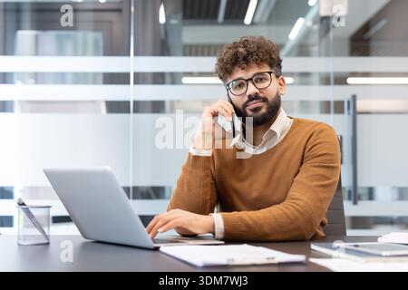 Junger Geschäftsmann mit lockigem Haar und Brille, der über sein Mobiltelefon kommuniziert, während er an einem modernen Arbeitsplatz an einem Laptop arbeitet und Geschäftsabläufe verwaltet Stockfoto