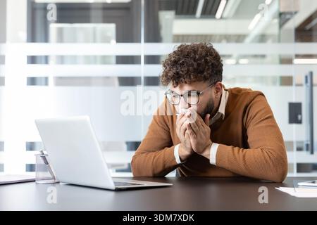 Junger männlicher Büroangestellter in braunem Pullover niest in ein Taschentuch an seinem Schreibtisch, während er einen Laptop benutzt, und zeigt Allergie- oder Erkältungssymptome während der Arbeitszeit in einem modernen Arbeitsplatz Stockfoto