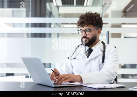 Junger männlicher Arzt, der an einem Laptop arbeitet, Online-Konsultationen anbietet und Patientenakten in einem modernen medizinischen Büro verwaltet, symbolisiert digitale Gesundheitsversorgung und professionelle Unterstützung Stockfoto