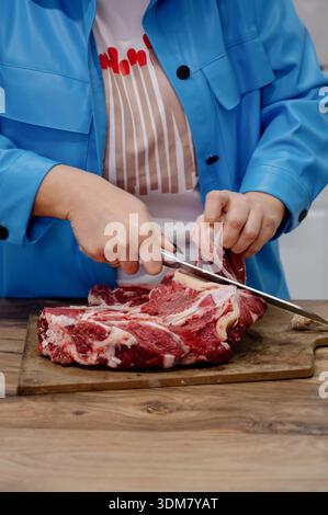 Das Schneiden von rotem Fleisch mit einem Messer auf einem hölzernen Küchentisch. Die Hände einer Frau schneiden frisches marmoriertes Rindfleisch auf einer hölzernen Tischfläche Stockfoto