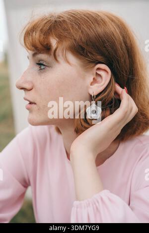 Nahaufnahme einer jungen Frau mit rotem Haar, heller Haut und blauen Augen, die eine rosa Bluse trägt und ihre Hand nahe ihrem Ohr hält. Sie sieht den an Stockfoto