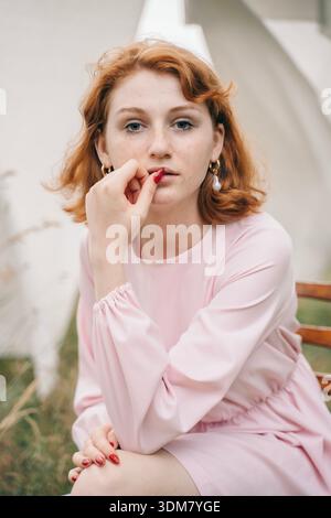 Porträt einer jungen rothaarigen Frau mit Sommersprossen in einem weichen rosa Kleid und goldenen Ohrringen, die auf einem Stuhl in einer natürlichen Umgebung im Freien sitzt. Sie l Stockfoto