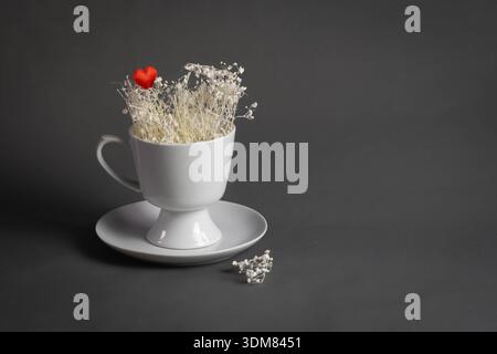 Romantisches Stillleben mit weißer Tasse, запечені-Blumen und rotem Herz auf dunklem Hintergrund Stockfoto