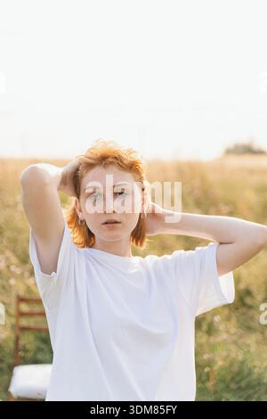 Natürliches Outdoor-Porträt einer rothaarigen Frau in einem weißen übergroßen T-Shirt, das auf einem sonnendurchfluteten Feld steht. Sie posiert selbstbewusst mit Händen hinter ihr er Stockfoto