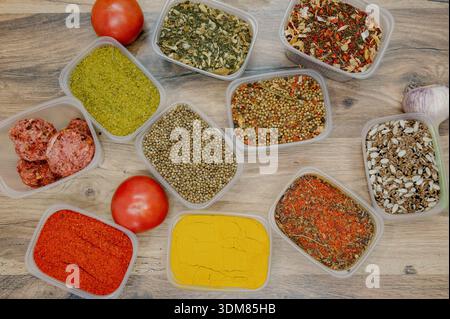 Eine Auswahl an gemahlenen und ganzen orientalischen Gewürzen mit einer roten Tomate auf einem Tisch. Draufsicht auf verschiedene getrocknete Gewürze und Gewürze in Kunststoffbehältern Stockfoto