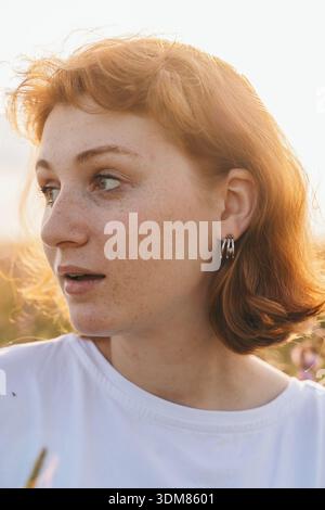 Nahaufnahme einer rothaarigen Frau, die zur Seite schaut, sanft beleuchtet durch Sonnenlicht der goldenen Stunde. Sie trägt ein weißes T-Shirt und minimalistische Creolen. T Stockfoto