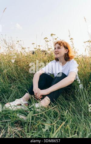 Eine fröhliche rothaarige Frau sitzt entspannt auf einer grünen Sommerwiese, trägt ein weißes T-Shirt, eine schwarze Hose und Creolen. Umgeben von Wildblumen Stockfoto