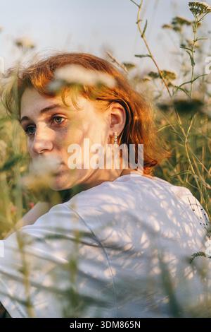 Eine rothaarige junge Frau mit Sommersprossen sitzt während der goldenen Stunde auf einer sonnendurchfluteten Wiese. Weiches natürliches Licht unterstreicht ihre zarten Merkmale, Silberschmuck Stockfoto