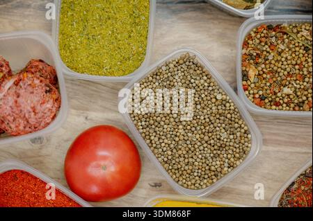 Eine Auswahl an gemahlenen und ganzen orientalischen Gewürzen mit einer roten Tomate auf einem Tisch. Draufsicht auf verschiedene getrocknete Gewürze und Gewürze in Kunststoffbehältern Stockfoto