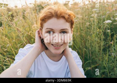 Porträt einer fröhlichen jungen Frau mit kurzen roten Haaren, silbernen Creolen und einem weißen T-Shirt. Sie sitzt auf einem Feld mit Wildblumen A Stockfoto