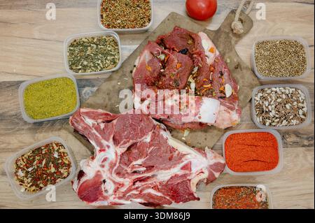 Rohe Rindfleischstücke auf dem Knochen mit Knoblauch und verschiedenen Gewürzen in Behältern auf dem Tisch. Frisches Rinderfilet auf einem Holzbrett umgeben von p Stockfoto