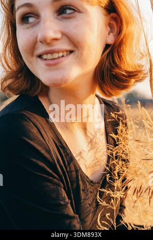 Porträt einer fröhlichen rothaarigen jungen Frau mit Sommersprossen, mit schwarzem Oberteil und zarter Halskette, die während des Sonnenuntergangs in einem Weizenfeld lächelt. Stockfoto