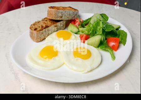 Frühstück mit Rührei mit Tomaten- und Gurkensalat auf geröstetem Vollkornbrot. Ein Teller mit Spiegeleiern, Gemüsesalat und knusprigem Baguette Stockfoto