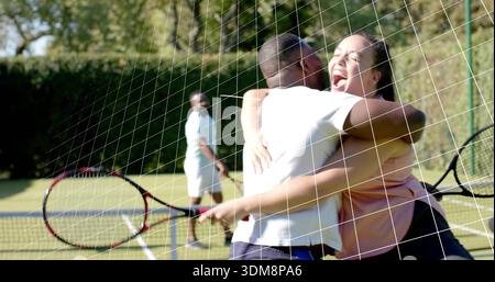 Umarmendes Paar, das in der Nähe des Netzes auf dem Rasenplatz feiert, Tennisschläger hält, Tenniskleidung trägt Stockfoto