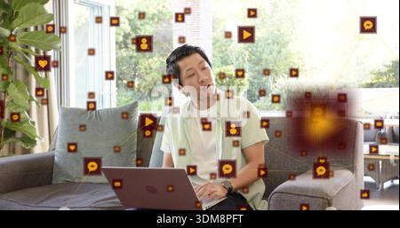 Asiatischer Mann in grünem Hemd auf grauem Sofa zu Hause, mit Laptop mit schwebenden Symbolen Stockfoto