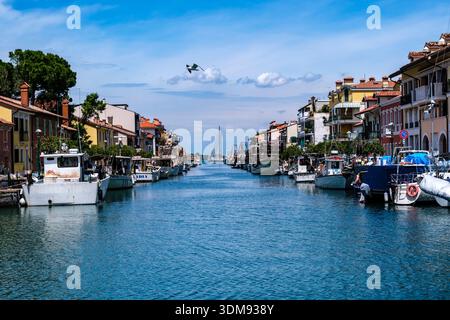 Blick entlang des Porto Canale in Grado mit vertäuten Booten und farbenfrohen Gebäuden unter einem blauen Himmel. Stockfoto