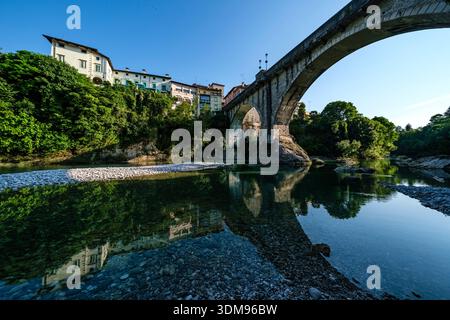 Die Steinbrücke Devil's Bridge überspannt den türkisfarbenen Natisone River mit historischen Bürgerhäusern von Cividale del Friuli in der Ferne. Stockfoto