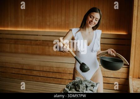 Eine Frau in einem stilvollen Badeanzug fügt heißem Wasser in einer ruhigen Sauna hinzu, um Entspannung und Wellness zu genießen. Stockfoto