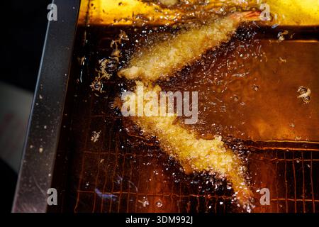 Zwei große panierte Garnelen, die in einem metallbehälter mit sprudelndem goldenem Öl in einer Küche frittiert werden. Stockfoto