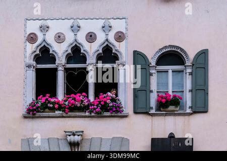 Detaillierter Blick auf die gotische Rathausfassade in Venzone mit verzierten Fenstern, rosa Geranien und Herzdekoration auf einer hellen Steinmauer. Stockfoto