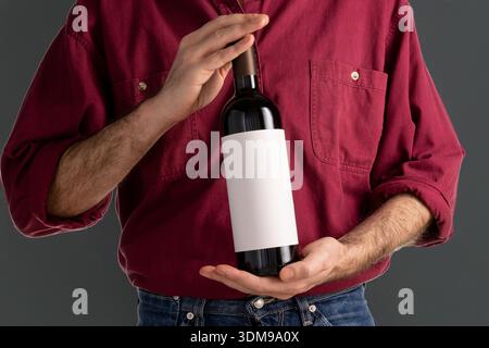 Der Typ im roten Hemd zeigt eine Rotweinflasche mit einem hochwertigen Papieretikett. Ideal für Modelle. Layout der grafischen Beschriftung. Online-Shop Stockfoto