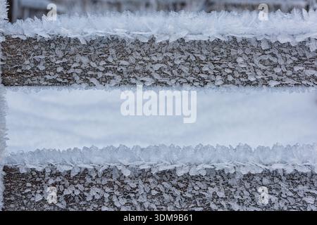 Ein Holzzaun mit zartem Frost fängt eine ruhige Winterszene ein, in der die Natur atemberaubende Muster vor weißem Schnee schafft. Stockfoto