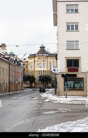 CELJE, SLOWENIEN, 01.03.2026: Eine ruhige urbane Straßenszene im Winter, mit einem historischen gelben Gebäude mit verzierten Fenstern, modernen Schaufensterfronten und einem Park Stockfoto