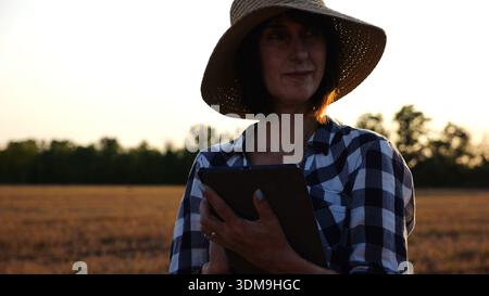 Agrostomistin im Strohhut, die die Ernte auf dem Gerstenfeld bei Sonnenuntergang überwacht. Erwachsener Landwirt mit digitaler Tablette auf Weizenwiese in der Abenddämmerung. Schön Stockfoto