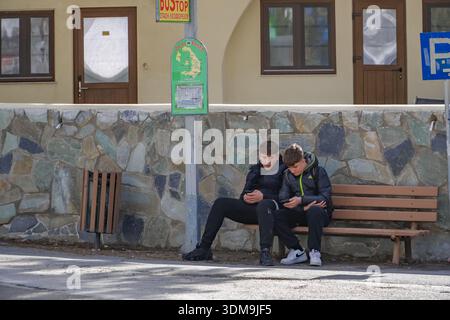 Junge Männer, die Handys benutzen, während sie an einer örtlichen Bushaltestelle in Emporio (Nimborios), Santorini, Griechenland, warten. Öffentliche Verkehrsmittel. Stockfoto