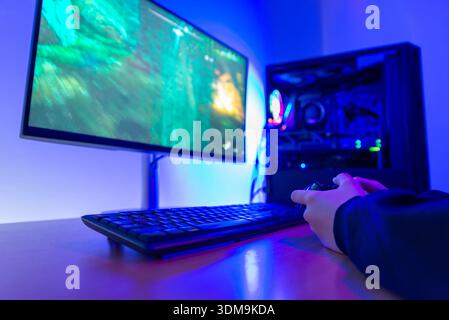 Gamer, der den Game-Controller hält, während er Videospiele auf dem Computermonitor an einem modernen Spieltisch mit beleuchtetem Desktop-pc spielt Stockfoto