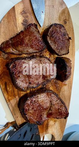 Perfekt gebratene, saftige mittelseltene Steaks ruhen auf einem rustikalen Holzschneidebrett mit einem scharfen Kochmesser. Eine köstliche, hochwertige kulinarische Szene. Stockfoto