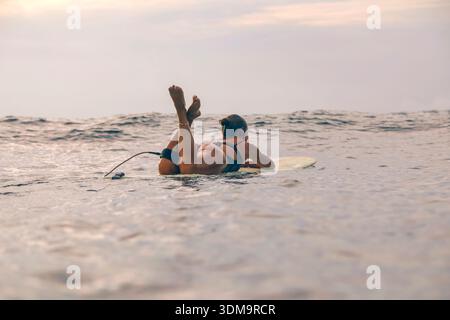 Junge Frau, Die Auf Dem Surfbrett Liegt Und Den Horizont Während Des Sonnenuntergangs Anstarrt, Entspannte Haltung Auf Sanftem Schwellen, Besinnlich Stockfoto
