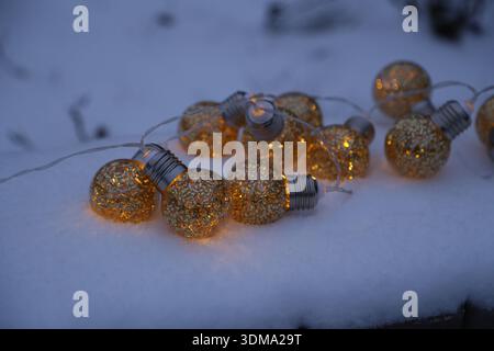 Lichterketten mit goldenen Glühbirnen werden auf einer Schneedecke platziert. Die Lichter leuchten auf und beleuchten den Bereich und erzeugen ein warmes Leuchten am kalten Abend. Stockfoto