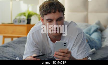 Ruhiger kaukasischer Mann liegend Bett zu Hause mit Smartphone bezahlen Online Kreditkartenkauf Zahlung fokussierte männliche Shopping Guy Käufer Kundenbank Stockfoto